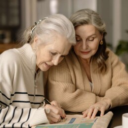 To ældre kvinder sidder sammen om en avis og skriver og lytter.