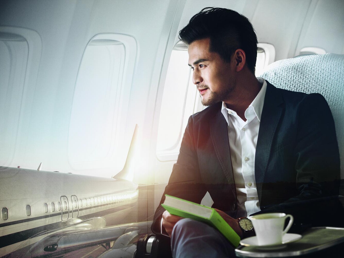 Billede af mand med ørepropper i ørerne i fly med grøn bog og en kaffekop.