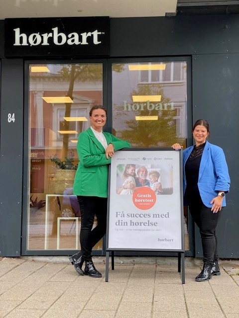 Indehaver Stinne Møller Nordby og medarbejder Tina Hillerup står foran Hørbarts klinik i Skanderborg ved et skilt med information om gratis høretest.